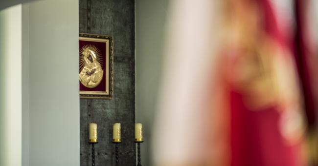 Fotografia wnętrza: złota ikona na ścianie, trzy świece; rozmyta flaga po prawej.