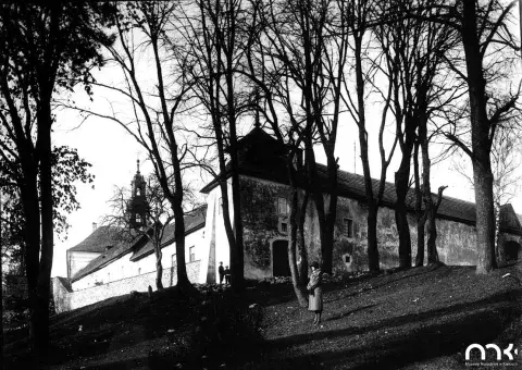 Czarno-biała fotografia: budynek na wzgórzu, nagie drzewa, dwie postacie