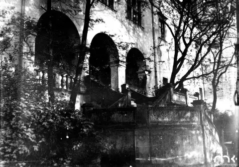 Fotografia czarno-biała: zaniedbane mauzoleum z łukami, nagrobki i omszałe drzewa