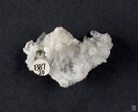 Fotografia białej kryształowej próbki minerału na czarnym tle z małą serduszkową etykietą.
