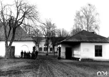 Czarno-biała fotografia wiejskiego dziedzińca z białymi budynkami, grupą ludzi i nagimi drzewami